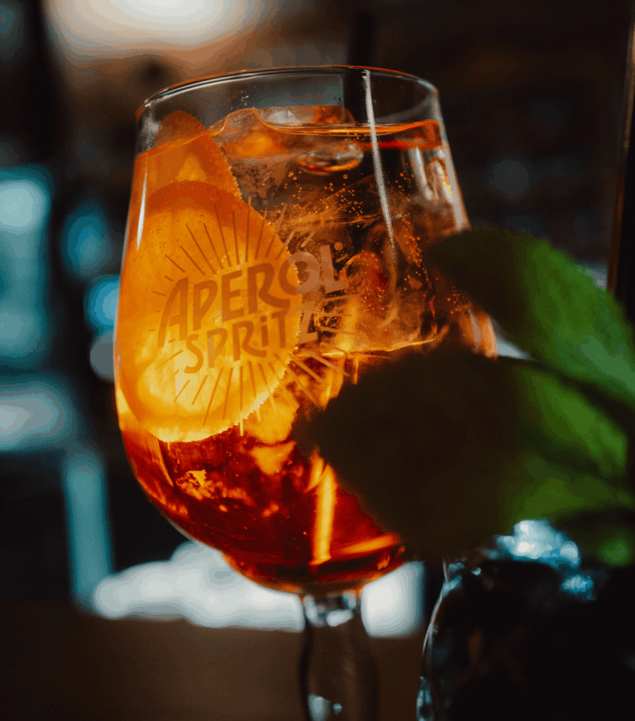 Nahaufnahme eines Aperol-Spritz-Glases mit Eiswürfeln, Orangenscheiben und frischer Minze, im Hintergrund unscharfes Barambiente.