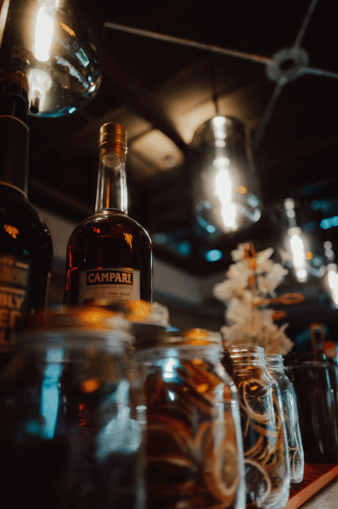Nahaufnahme einer Bar mit dekorativer Campari-Flasche, Glasbehältern mit getrockneten Zitrusschalen und moderner Hängelampe im Hintergrund.
